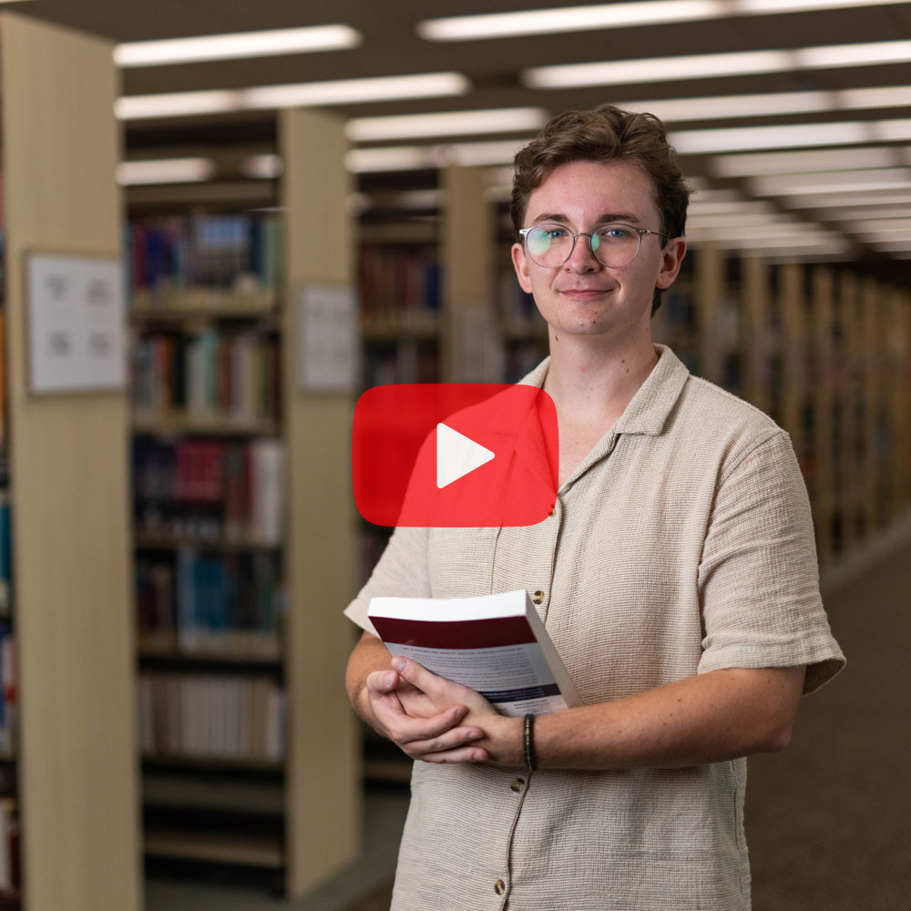 A male student wearing a beige shirt holding textbooks smiling to camera in the library