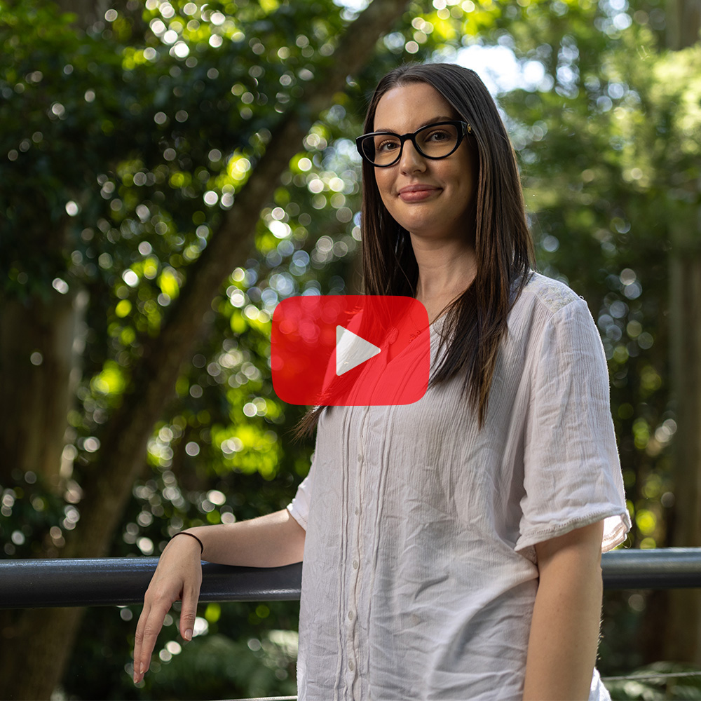 A female student wearing a white shirt and glasses smiling to camera standing in front of bushland