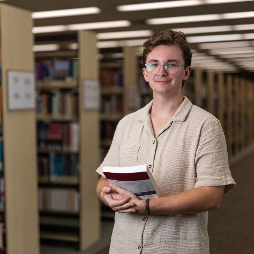 A student standing in the library holding textbooks smiling to camera