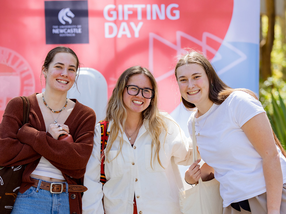 Three students smiling to camera standing in front of a red media wall