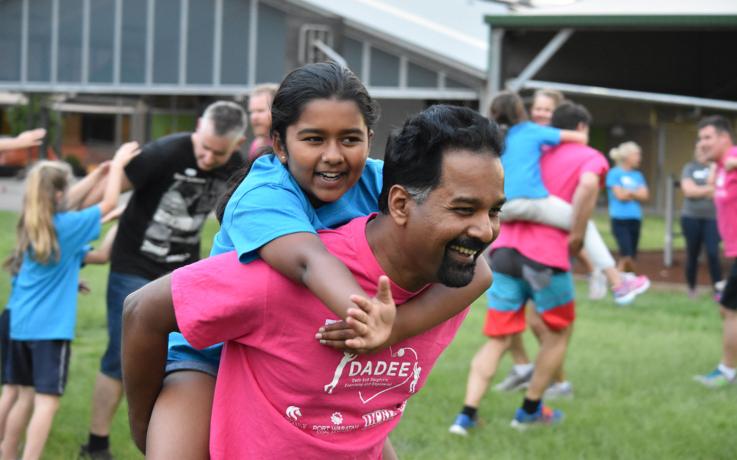 A father giving his daughter a piggy back ride with other dads and daughters in the background A father giving his daughter a piggy back ride with other dads and daughters in the background