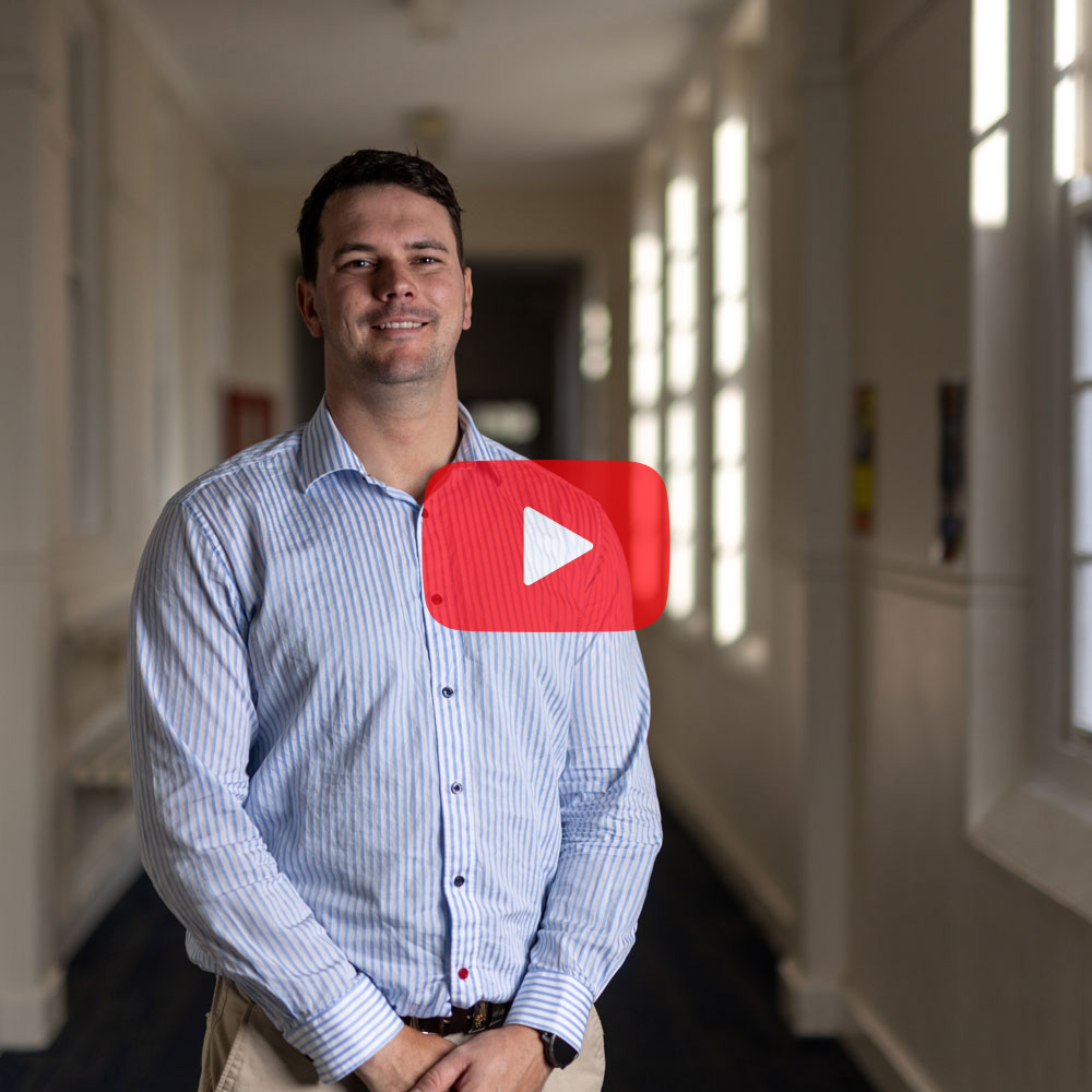 A male student wearing a blue shirt standing in a school hallway smiling to camera